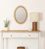 A gold oval mirror hangs on a white wall above a white console table with a wooden top. A lamp, a plant, and a candle are on the table. Náhľad obrázku produktu