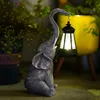 A solar-powered elephant statue holding a lantern in a garden setting. Produktbild-Vorschau