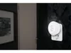 A white LED night light is plugged into a wall socket on a dark patterned wallpaper. A framed picture is visible on the left side of the image. Produktbild-Vorschau