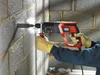 A person wearing gloves is using a Black & Decker rotary hammer drill to drill into a concrete block wall. Dust is visible around the drill bit. Produktbild-Vorschau