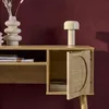 A close-up view of a wooden desk with a rattan-inlaid door, a lamp, and decorative vases against a dark red wall. Produktbild-Vorschau