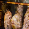 Several rolls of meat wrapped in netting are hanging from a wooden rod inside a smoker or curing chamber. Produktbild-Vorschau