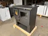 A dark gray wood-burning stove is displayed on a wooden pallet in what appears to be a warehouse or storage area. Produktbild-Vorschau