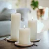 Three white LED pillar candles with lit flames are arranged on a wooden surface, with a blurred background of a living room. Produktbild-Vorschau