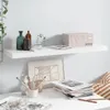A white floating shelf is mounted on a light gray wall above a white desk. The shelf holds decorative items, including bottles, a plant in a glass vase, and a box. The desk has art supplies, magazines, and a framed picture. Náhľad obrázku produktu