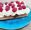 A close-up of a waffle topped with whipped cream, raspberries, and bananas, served on a blue plate. Anteprima dell’immagine prodotto