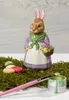 A ceramic Easter bunny figurine holding a basket of eggs, surrounded by moss, painted eggs, and a paintbrush with a can of green paint. Náhled obrázku produktu