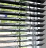 Close-up view of black blinds with a window and greenery visible through the slats. Produktbild-Vorschau