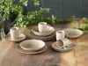 A set of stoneware dishes and mugs arranged on a wooden table with greenery in the background. Produktbild-Vorschau