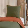 A close-up view of a bed with green bedding and an orange throw blanket, set against a wooden headboard and a decorative lamp. Produktbild-Vorschau