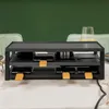 A black raclette grill is set up on a table with a white tablecloth. In the background, there are two chairs and a green wall. Produktbild-Vorschau