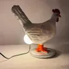 A white chicken-shaped lamp with orange feet and a red comb sits on a round base, plugged into a power cord. Podgląd zdjęcia produktu