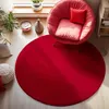 A red circular rug is laid out on a light wooden floor, with a red accent chair and a white knitted pouf placed on it. Plants and a small bowl are visible on a windowsill in the background.