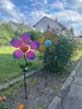 Two solar-powered garden stakes shaped like flowers are planted in a garden bed next to a sidewalk. Produktbild-Vorschau