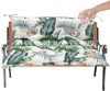 A hand is pouring water onto a bench cushion with a tropical leaf pattern, demonstrating its water-repellent feature. Produktbild-Vorschau