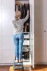 A woman stands on a stool in front of an open closet, reaching for clothes. Náhled obrázku produktu