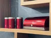 A red bread box and three red canisters are on a wooden shelf in front of a dark marble backsplash. Náhled obrázku produktu