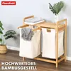 A bamboo laundry sorter with two beige laundry bags, placed in a room with a plant and folded towels on top. Podgląd zdjęcia produktu
