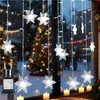 Snowflake-shaped lights are hung on a window, with a Christmas tree and falling snow visible in the background. Anteprima dell’immagine prodotto