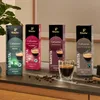 Four boxes of Tchibo Cafissimo coffee capsules are displayed on a dark surface, with a shot of espresso in a glass in the foreground and coffee beans scattered around. Náhled obrázku produktu