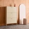 A cream-colored metal shoe cabinet with three drawers is shown against a wooden wall. A decorative vase and a striped stool are also present. Produktbild-Vorschau