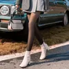 Person in schwarzer blickdichter Strumpfhose, weißen Stiefeletten, grauem Rock und kariertem Blazer, mit grauer Handtasche, an einem grünen Auto vorbeigehend Anteprima dell’immagine prodotto