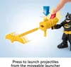 A close-up of a Batman toy figure holding a yellow launcher with a red projectile being inserted by a finger. The launcher is extended and has a bat-shaped projectile at the end. Text at the bottom explains how to launch projectiles. Podgląd zdjęcia produktu