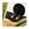 A pair of black wireless earbuds with wood accents are shown in their open charging case, resting on a wooden surface with a blurred green plant in the foreground. Produktbild-Vorschau