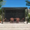 A bioklimatische pergola with a table and chairs set up on a wooden deck, surrounded by greenery and a stone wall. Produktbild-Vorschau