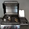 A stainless steel grill with the lid open, showing the cooking grates and accessories. Podgląd zdjęcia produktu