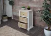 A white and natural wood bedside table with rattan drawer fronts, placed on a wooden floor next to a rug and a potted plant. Anteprima dell’immagine prodotto