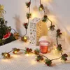 A festive Christmas decoration featuring a string of LED lights intertwined with pine sprigs, pinecones, and red berries, arranged on a textured surface next to a small Christmas tree and decorative lanterns. Produktbild-Vorschau