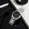 A close-up shot of a men's watch on a reflective black surface, with a pen and a notebook to the left, and a marble-like surface to the top right. Produktbild-Vorschau