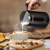 A hand pours frothed milk from a black milk frother into a white cup of coffee, creating latte art. There is a pastry on a plate to the left. The scene is set on a wooden board with coffee beans scattered around. Produktbild-Vorschau