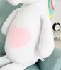 Close-up of a large white plush unicorn toy with a pink heart on its belly, sitting on a light blue couch. A black alarm clock is visible in the background. Podgląd zdjęcia produktu