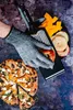 A hand wearing a protective glove is using a grater to slice an apple, with sliced apples and pumpkin on a plate nearby, and a finished pizza-like dish in the foreground. Produktbild-Vorschau