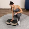 A woman is kneeling on a rug next to a vibration plate, holding a remote control. Náhled obrázku produktu