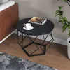 A round black coffee table with a geometric metal base is placed on a wooden floor next to a sofa and a potted plant. Produktbild-Vorschau