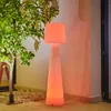 A tall, illuminated floor lamp with a pleated shade stands next to a small tree on a patio. Produktbild-Vorschau