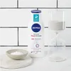 A Nivea body lotion bottle is displayed next to an hourglass and a small bowl of cream, set against a tiled background. Voir les images du produit
