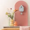 A modern cuckoo clock is displayed on a pink arch background, next to a vase of flowers and decorative glass objects. Náhled obrázku produktu