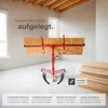 A red drywall lift holding a large sheet of particle board in a room with wooden beams on the ceiling and windows in the background. There are stacks of particle board on the right side of the image. Náhled obrázku produktu