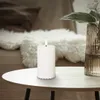 A white LED candle with a flame sits on a round wooden table in a cozy room setting. Podgląd zdjęcia produktu