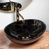 A black marble basin with gold accents and a gold faucet is shown on a wooden surface, with a gold-rimmed mirror in the background. Produktbild-Vorschau