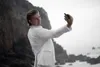 A man in a white suit takes a selfie on a rocky beach. Voir les images du produit