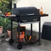 A black Royal Gourmet charcoal grill is set up on a wooden deck, with plants and a storage bin in the background. A bag of charcoal and a small bag are on the lower shelf. Produktbild-Vorschau