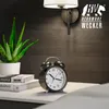 A close-up of a black analog alarm clock on a white nightstand next to a potted aloe vera plant and some books, with a wall lamp in the background. Anteprima dell’immagine prodotto