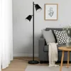 A modern living room scene featuring a black floor lamp with two adjustable heads, a grey sofa with decorative pillows and a throw blanket, and a small wooden side table. Náhled obrázku produktu