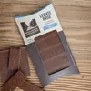 A chocolate bar in its packaging, with a few broken pieces of chocolate scattered around it, placed on a wooden surface. Produktbild-Vorschau