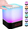 A hand is touching the top of a cube-shaped bedside lamp that also functions as a Bluetooth speaker and alarm clock. The lamp displays a gradient of colors, and a digital clock is visible on the front. Produktbild-Vorschau
